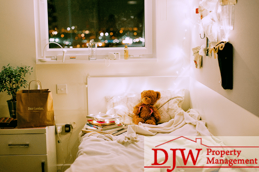 A bed in the corner of the room with white sheets, a teddy bear, and books. There are decorations on the wall and a small window behind the bed.